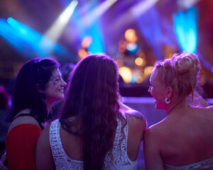 Three Female Friends Watching Band Dancing At Outdoor Summer Music Festival Holding Drinks At Night