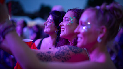 Three Female Friends Watching Band Dancing At Outdoor Summer Music Festival Holding Drinks At Night