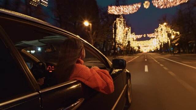 Young smiling woman in the front seat of the car looking out of the window with amazement of how beautiful is the city street with Christmas lights at night. Winter Christmas season