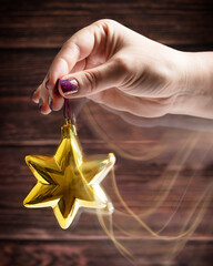 Palm of female hand with bright lilac manicure holding shiny star shaped christmas bauble of gold colour with blurred motion against dark brown wooden background used for winter holiday greetings