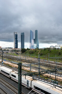 Elevated panorama of several train tracks and a moving train with four modern high-rise office towers in the background in the city of Madrid in Spain