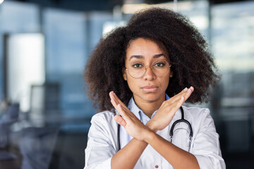 Close-up photo. Portrait of a young African American female doctor standing in the office of the clinic, looking seriously at the camera, crossing arms and showing a sign of prohibition and protest - obrazy, fototapety, plakaty