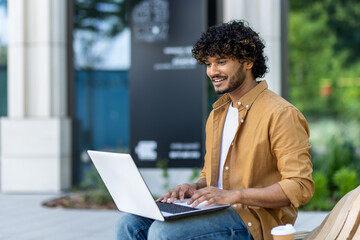Indian young man sitting on the street with a laptop and texting, talking on a video call with a...