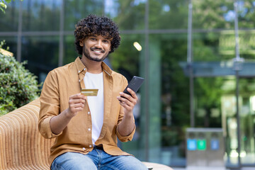 Portrait of a smiling young Indian man sitting outside on a bench, holding a credit card and a...