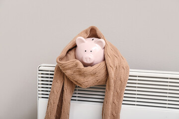 Piggy bank with scarf on radiator near light wall. Winter heating concept