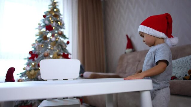 Caucasian toddler in grey pajama and red Santa cap stands near the desk. Kid leans on the desk and raises his feet. Christmas tree at backdrop in blur.