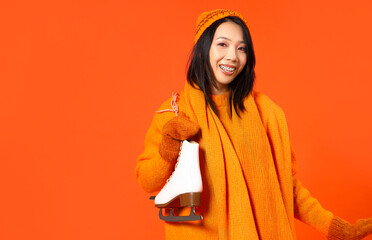 Young Asian woman in winter clothes with ice skates on orange background