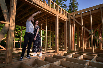 Man and woman inspecting their future wooden frame house nestled in the mountains near forest....