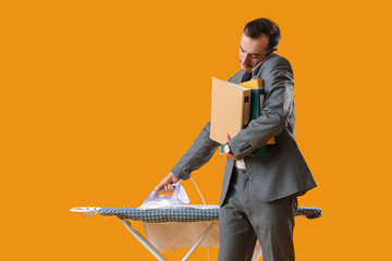 Young businessman with folders ironing shirt on board against yellow background