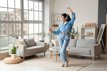 Young African-American woman in headphones with mobile phone dancing at home