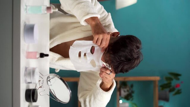 Young arab men wears bathrobe applying face sheet mask, looking at the mirror and doing daily morning beauty routine.
