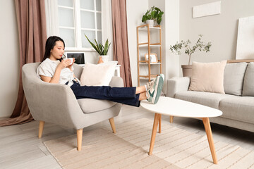 Young Asian woman drinking coffee in armchair at home