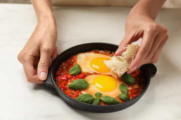 Woman dipping piece of bread into delicious Shakshuka at white table, closeup
