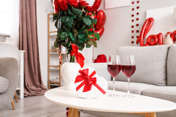 Vase with roses, gift and glasses of wine on table in living room decorated for Valentine's Day