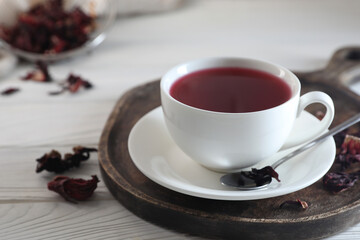 Delicious hibiscus tea in cup and dry roselle petals on white wooden table, closeup