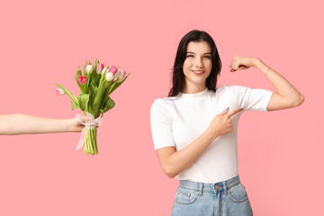 Young woman ignoring bouquet of flowers and flexing muscles on pink background. Feminism concept