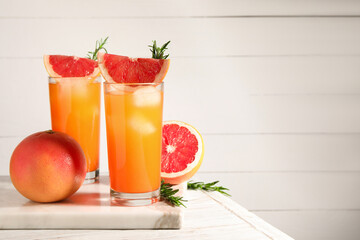 Tasty grapefruit drink with ice in glasses, rosemary and fresh fruits on light wooden table. Space for text