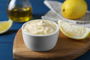 Tasty mayonnaise in bowl and lemon wedge on blue table, closeup