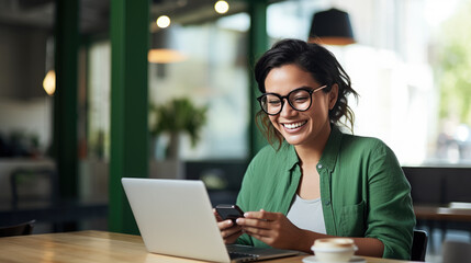 Cheerful professional woman wearing glasses and a green blouse is sitting at a desk with a laptop and holding coffee cup - obrazy, fototapety, plakaty