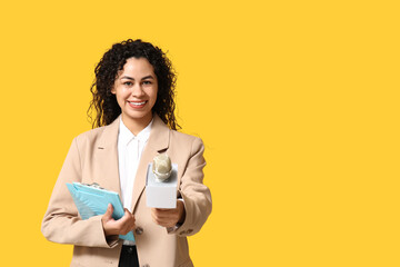 Beautiful young happy African-American female journalist with microphone and clipboard on yellow background