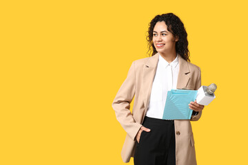 Beautiful young happy African-American female journalist with microphone and clipboard on yellow background