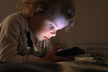 Beautiful cute adorable blonde wavy haired baby girl using smart phone before sleeping looking at...