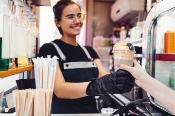 Smiling young barista girl in casual clothes, apron and black gloves gives cocktail with cream and...