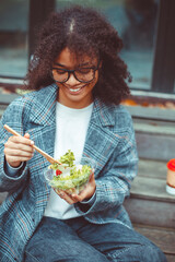 Happy african american woman office worker eating salad outdoors while sitting on bench in park