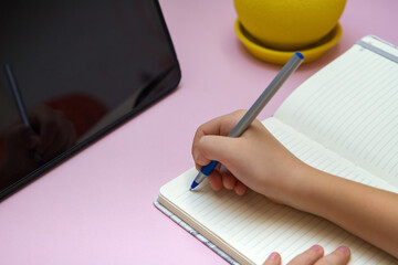 Child's hand writing in notebook on pink background. Education concept.