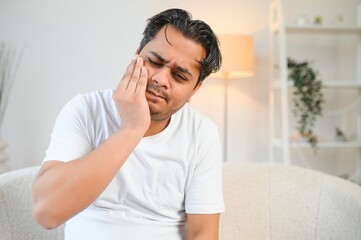 Dental problems. Young indian man touching cheek, closing eyes with expression of terrible suffer from painful toothache, sensitive teeth, cavities. Hindu guy at home apartment living room on sofa