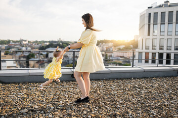 Beautiful smiling brunette holding little daughter in arms and spinning on roof of high-rise...