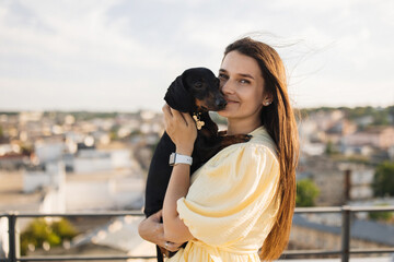 Portrait of cheerful woman with long dark hair standing on rooftop and embracing black dog in arms. Caucasian young female walking with lovely pet during warm summer day.
