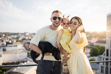 Happy adult parents wearing stylish sunglasses standing on balcony of high-rise building and...