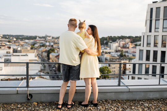 Positive married couple with little daughter embracing on gravel rooftop with beautiful cityscape and blue sky. Cheerful family holding each other tightly and spending time on fresh air.