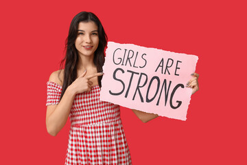 Young woman pointing at sign with text GIRLS ARE STRONG on red background. Feminism concept
