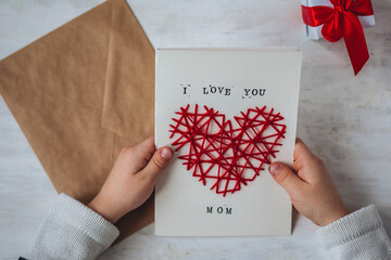 Concept of children craft, handmade gift for Mother's Day. Little boy holding craft card with embroidering in the shape of heart and printed message I love you Mom. Kid's leisure activity, handcraft - obrazy, fototapety, plakaty