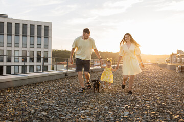 Front view of friendly people dressed in bright clothes walking on rooftop at sunset. Caucasian family of father, mother, little daughter and dachshund dog holding hands and looking at horizon. - Powered by Adobe