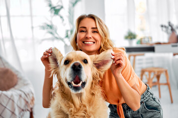 Fun with animal. Carefree woman joyfully lifting ears of adorable golden retriever and smiling at camera while sitting on floor. Young pet owner enjoying every moment spending with her best companion. - obrazy, fototapety, plakaty