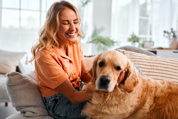 Love for animals. A cheerful happy woman with wavy blonde hair is petting a golden retriever while relaxing on the sofa.