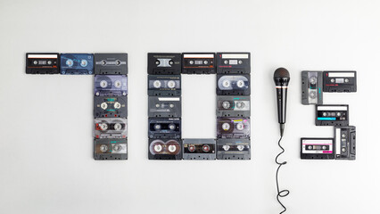 The text "70's" on a gray paper background, composed of ancient, different, colorful audio cassettes and mic. The year of nostalgia for karaoke parties 1970's