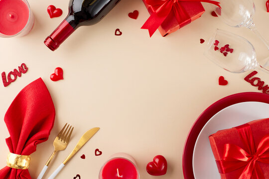 Sharing a candlelit dinner on Valentine's Day. Top view photo of plates, cutlery, wine bottle, wineglasses, red hearts, napkin, gift box, candle on beige background with promo space