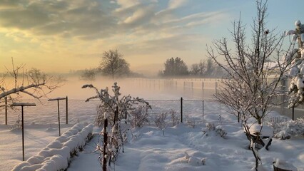 Moving fog with orange sunlight during sunrise over fields with snow behind garden with fence near noise barriers along national road in Poland with horizon with trees - timelapse. - Powered by Adobe