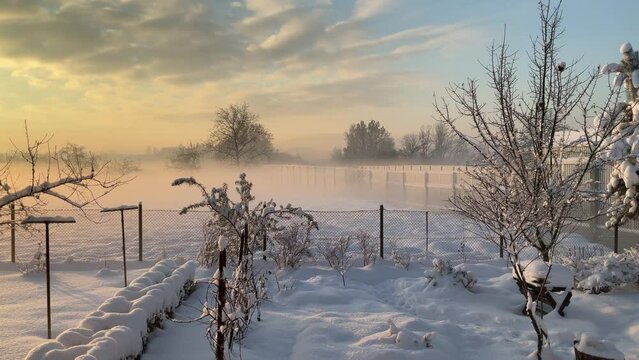 Moving fog with orange sunlight during sunrise over fields with snow behind garden with fence near noise barriers along national road in Poland with horizon with trees - timelapse.