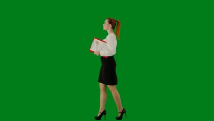 Fototapeta na wymiar Portrait of attractive office girl on chroma key green screen. Woman in skirt and blouse walking holding red folder. Side view.