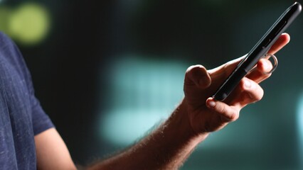 Young man walking around town at nighttime, using mobile phone under streetlamps strolling near urban buildings. Person enjoying walk texting messages on sidewalk. Handheld shot. Close up.