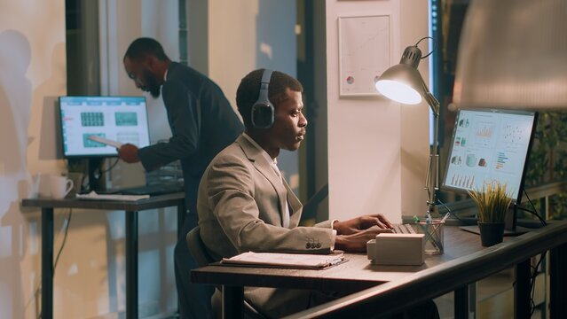 Employee enjoying relaxing music to take his mind away from tedious repetitive project tasks during nightshift. Accountant in office with headphones on checking audit figures and diagrams