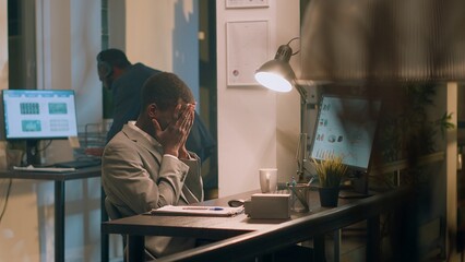 African american accountant exasperated with noisy colleague listening to music and pretending to beat drums during nightshift. Employee annoyed by distracting coworker banging desk in office