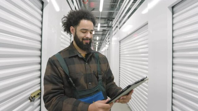 Smiling young man with stylish afro curls in a long hallway with illumination clicking on tablet standing in front of white metal roller shutter doors. High quality 4k footage