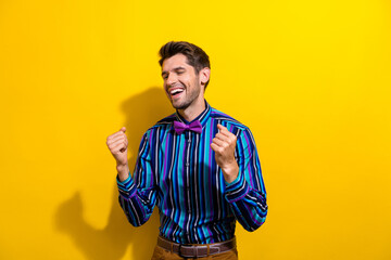 Portrait of optimistic overjoyed guy dressed stylish shirt bow tie eyes closed clenching fists dancing isolated on yellow color background