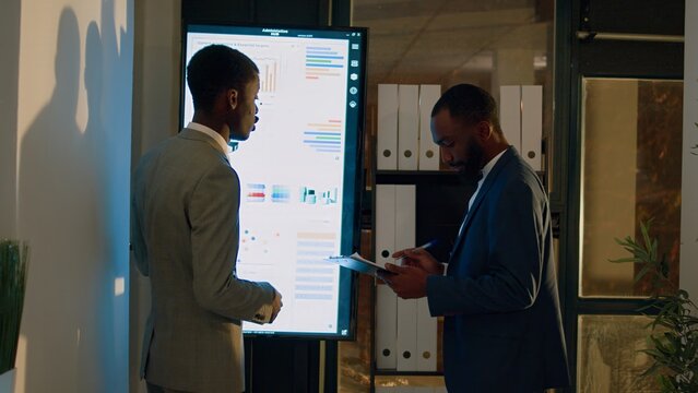 Manager debriefing employee in front of digital screen, showing him important research statistics before getting back to work. Businessmen in office during nightshift brainstorming business insights